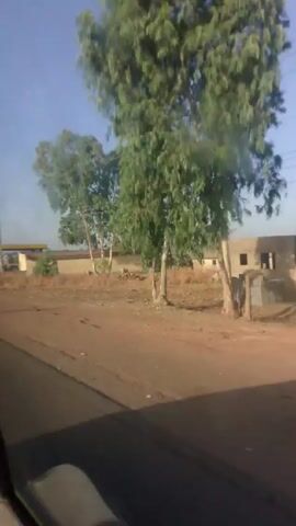 The video shows a view from inside a moving car driving down a paved road on a clear, sunny day. The road is dry and dusty, and the landscape appears to be rural, with dry vegetation and sandy areas alongside the road. In the distance, there are buildings and telephone poles visible. A gas station can be seen on the side of the road. As the car passes the gas station, people and vehicles are visible, including a motorcyclist. The camera appears to be mounted inside the car, capturing the perspective of someone in the driver's seat or passenger seat. The general impression is that the video was taken in a developing area, likely in a hot and dry climate.
