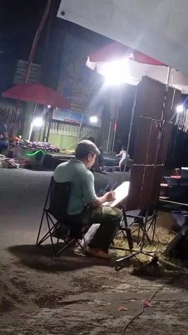 A man wearing a hat and a teal shirt sits in a foldable chair and draws on a pad. He is outdoors at night, with artificial lights illuminating the scene. To his left, there are stalls with items displayed, suggesting a marketplace or a street fair. To his right, there are wooden panels and more lighting equipment, possibly for a performance or display. Another person is visible further in the background, also illuminated by the lights. The ground is paved with asphalt and dirt.