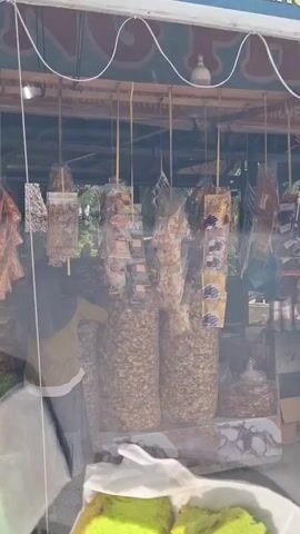 This video shows a market stall selling various snacks. The stall is covered by a canopy, and various items are displayed, including large bags of snacks hanging from the ceiling. A woman wearing a headscarf and a long skirt is browsing the items. In the foreground, two pieces of green cake are visible on a white plate. The overall impression is of a bustling outdoor market scene.