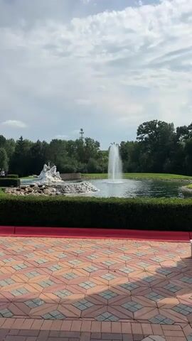 A wide shot captures a serene outdoor scene with a pond in the foreground. A large fountain sprays water high into the air in the center of the pond. To the left of the fountain, a white sculpture of multiple figures sits on a tiered rock structure. A neatly trimmed hedge runs along the edge of the pond, separating it from a paved area with a decorative, brick-like pattern. Beyond the pond, a line of lush green trees forms a backdrop, and a tall, utilitarian tower with antennas is visible in the distance against a partly cloudy sky. The image is stable, indicating it was likely filmed from a stationary position.