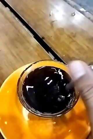 The camera shows a clear glass cup filled with black liquid sitting on a bright yellow saucer. A metal spoon rests inside the cup. The cup is on a wooden table with a black stripe running across it.