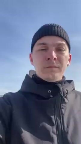 The video shows a man's face looking directly at the camera. He is wearing a black beanie and a black hooded jacket. He appears to be outside on a sunny day with a clear blue sky in the background. A patch of snow and a building are visible in the background. The camera is positioned slightly below him, looking up. The man is not visibly interacting with anything or anyone.