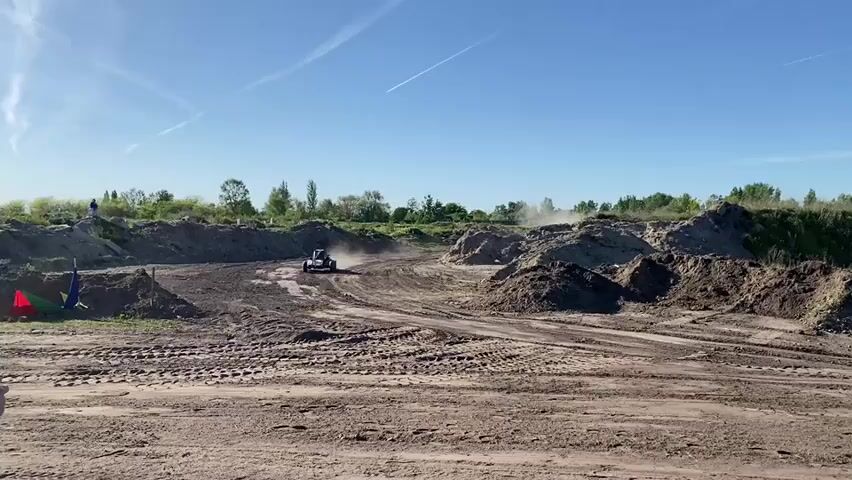 The video shows a blue and silver off-road buggy driving on a dirt track. The buggy kicks up dust as it navigates around dirt mounds. In the background, there's a grassy field with vehicles parked, including a van and a yellow construction vehicle. A traffic light stands on a pole, and there are houses in the distance. The sky is clear and blue with contrails. The track appears to be prepared for racing or off-road driving.