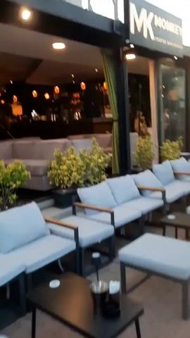 A video pans from left to right, showing the exterior of a lounge bar and hookah establishment called "MK MONKEY LOUNGE BAR/HOOKAH". In the foreground, there are several outdoor seating areas with light blue cushioned sofas and low tables. Green potted plants are strategically placed around the seating areas. The entrance to the establishment features a bar area with patrons visible inside. The exterior walls of the building are black, and a neon sign displays the establishment's name. The video ends with a shot of an area with green artificial turf and a woman walking by in the foreground.