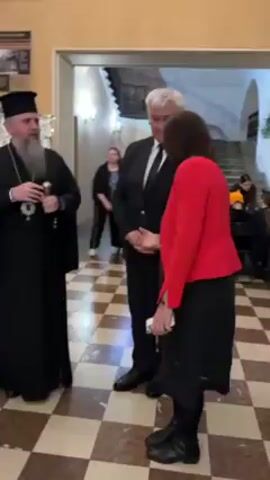 The video shows three people standing in an ornate room with a checkered tile floor. The person on the left is a man wearing a black robe and a black hat, with a long, gray beard. The person in the middle is a man wearing a black suit and a white shirt with a black tie. The person on the right is a woman wearing a red jacket over a red and black dress. The man in the black suit is smiling and looking at the woman in the red jacket. The man in the black robe is smiling and looking toward the camera. They appear to be posing for a photo, though no camera is explicitly visible. There are other people in the background, as well as a staircase and some artwork on the wall.