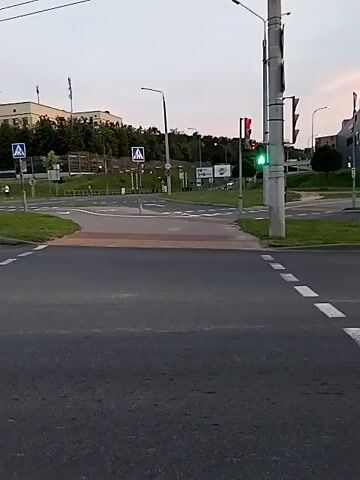 This is a video of a street intersection. The traffic light is red for the direction of the camera. There are crosswalks and sidewalks visible. In the background, there are buildings and trees. The sky is overcast.