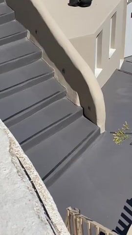 The video begins by focusing on a dark gray staircase and the light-colored walls and ground surrounding it. A small wooden fence is visible in the foreground. The camera then pans upwards and to the right, revealing more of the light-colored architecture, including a white door. Green plants and cacti are strategically placed around the property. As the camera continues to move, a glimpse of a swimming pool with bright blue water is seen. The perspective shifts to show a wider view of the coastline with white buildings perched on a rocky cliff overlooking a deep blue sea. A boat is seen moving across the water in the distance. The final shot offers a panoramic view of the sea, the cliffside village, and the vast ocean under a clear sky.