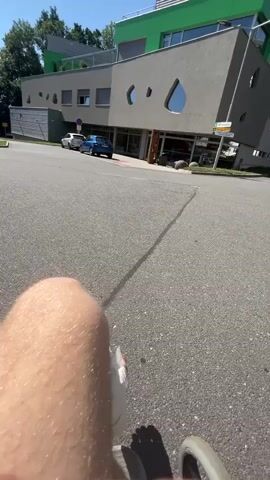The video shows a first-person perspective of someone in a wheelchair, moving forward on an asphalt path. The scene is outdoors on a sunny day, with clear blue skies and bright sunlight casting shadows.

In the foreground, a person's bare leg is visible, with a bandage wrapped around the knee. The wheelchair's wheel is partially visible at the bottom right of the frame.

In the background, there is a modern building with a grey facade and a distinctive green upper story. The building has unique, irregularly shaped windows. To the left of this building, there are other buildings and trees. Two cars are parked in front of the grey building: a white car and a blue car.

The location appears to be an outdoor area adjacent to a building, possibly a hospital or a similar institution given the modern architecture and the presence of a person with a bandaged knee in a wheelchair. Signage is visible in the background, including a yellow sign with an arrow and some text, and a white sign with text and graphics. No readable text is clearly visible. The overall mood is one of movement and recovery, under a bright, warm day.