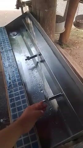 A first-person perspective shot shows a hand reaching for a faucet attached to a metal trough. Water flows from the faucet into the trough, creating ripples and splashes. The trough is long and rectangular, with a tiled border along the left side. Two metal spigots are positioned along the center of the trough. In the background, a wooden post supports a structure and a mesh bin is visible. The ground appears to be dirt or gravel.