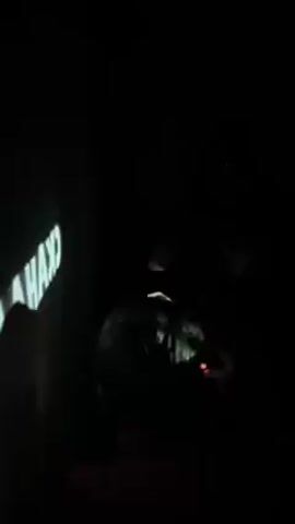 A dark room shows two people, one in the foreground and one in the background. The person in the background is wearing dark clothing, sunglasses, and holding up two fingers on each hand. A projector displays images on a wall to the left of the screen. The person in the foreground is facing away from the camera and looking at a small screen. The room is dark and difficult to make out details.