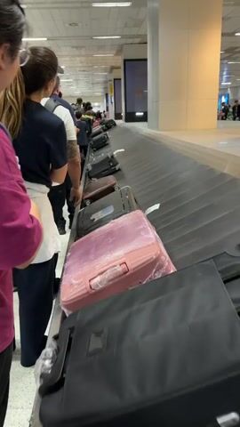 The video shows a luggage carousel at an airport. Several pieces of luggage, including a pink suitcase wrapped in plastic, are moving along the carousel. A line of people stands to the left of the carousel, waiting to collect their bags. The airport environment appears busy, with bright lighting and digital displays visible in the background. The camera is positioned at eye level with the carousel, focusing on the luggage and the people waiting.
