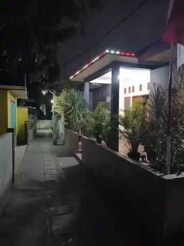 This is a short video clip filmed at night, showing a narrow alleyway next to some houses. The alleyway is paved with flagstones and is dimly lit. On the right side of the alley, there is a raised planter with several green plants. Behind the planter is a house with its porch light on, illuminating the entrance. The house has a dark-colored roof with decorative white and red lights along the edge. A flag pole with a flag is visible on the right side of the house. On the left side of the alley, there is a yellow house with a blue door. Trees and more houses are visible in the background, also shrouded in darkness. The video is shaky and appears to be handheld. There is no discernible sound other than a voice speaking in what sounds like Indonesian.