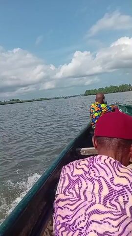 Here's a concise description of the video:

Three people are riding in a motorboat on a river or large body of water. The person closest to the camera is wearing a purple patterned shirt and a red cap. Another person is wearing a brightly colored patterned shirt. The third person is wearing a dark dress and a straw hat. The boat has some writing on the side. The sky is blue with scattered white clouds, and the shoreline is visible in the distance, with trees and some buildings. The boat is moving, creating ripples in the water.