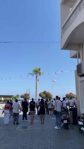A vertical, eye-level shot captures a bright, sunny day outdoors. In the foreground, a diverse group of people, viewed from behind, walk along a paved walkway lined with what appears to be stone tiles. They are all facing away from the camera and walking towards a palm tree in the distance. A white moped is parked on the right side of the frame. The middle ground features the tall, slender trunk of a palm tree adorned with a Turkish flag on its left and another flag, red and white, on a pole to its right. A string of small red and white triangular flags hangs in an arc above them. Behind the palm tree, there's a large, rounded shrub with red flowers. More people are visible in the background, gathered and looking towards something out of frame. A glimpse of the sea can be seen beyond them, with a distant structure on the horizon. The background is dominated by a clear, bright blue sky. On the right side of the frame, a white building with arched windows and an air conditioning unit is partially visible. The overall atmosphere suggests a public gathering or a leisurely stroll on a warm day.
