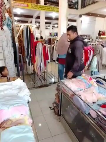 The video shows the interior of a clothing store. In the foreground, on the left, there are racks of folded clothes, mostly light-colored with pink and yellow accents, displayed on a table. To the right of this, a glass-topped display case contains more folded clothing, also in pastel colors like pink and blue. In the mid-ground, a man in a dark jacket and blue jeans stands facing away from the camera, looking towards a rack of clothes. He is positioned to the right of center. To his right, a woman wearing a light blue headscarf and a light blue shirt, also facing away from the camera, appears to be browsing the clothing racks. Behind them, more clothing racks are visible, displaying various garments in colors such as red, white, and cream. The store appears to have a tiled floor and a high ceiling with visible support beams and lighting fixtures. A sign with Indonesian text is visible on the wall in the background. On the far left, a child in a black shirt is sitting on the floor, facing the clothing racks. The overall impression is a bustling retail environment with people shopping for clothes.