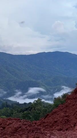 The video opens with a wide shot of a mountainous landscape. The mountains are covered in lush green trees, and a layer of white clouds or fog is visible in the valleys between them. The sky is overcast with a mix of light and dark clouds. In the foreground, on the right side of the frame, there is a mound of reddish-brown soil. The camera then pans upwards, focusing on the sky. The clouds become more prominent, with some appearing dark and heavy, suggesting the possibility of rain. A thin black wire is visible diagonally across the frame in the upper portion, stretching from left to right. The video continues to focus on the sky and clouds, with the landscape gradually disappearing from view as the camera's angle shifts.