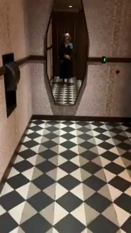 A short video shows a woman taking a mirror selfie in a bathroom with a black and white checkered floor and patterned wallpaper. The woman is reflected in a large, irregularly shaped mirror. She is wearing a teal dress, carrying a tan purse with a chain, and wearing bright pink shoes. She lifts her hand to her curly hair. The bathroom has a wooden wall and a door visible in the mirror's reflection. A black paper towel dispenser is mounted on the wall next to the mirror.