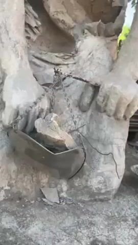 The video shows a cement sculpture of a woman holding scales. The sculpture appears weathered and worn, with cracks and missing pieces. The woman is dressed in what appears to be a ragged cloth. The scales hang from chains and are filled with rocks. A black car is visible in the background. The scene is outdoors, likely in a park or garden setting.