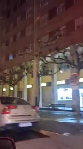 The video shows a nighttime scene from a moving car in an urban environment. A tall, brown brick building with many windows dominates the background.  Trees with bare branches line the street, with streetlights casting a warm glow. Several cars are visible, including a white sedan, a white SUV, and a silver hatchback, all traveling in the same direction. The overall atmosphere is typical of a city street at night.