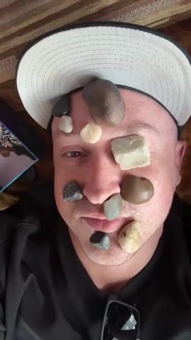 A man wearing a black shirt and a white baseball cap lies on his back on a patterned surface. He has several stones placed on his face and head. His eyes are open and he is looking at the camera. A person's hands enter the frame and are seen touching the man's face and removing stones. The person speaking in the background expresses disapproval of the action.