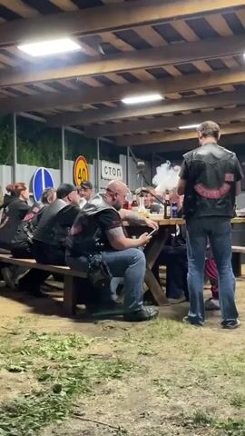 Several people are seated at a picnic table under a covered outdoor structure. They appear to be part of a motorcycle club, as they are wearing leather vests with the club logo on the back. Some are drinking beer, while one person is looking at their phone. Smoke is wafting through the air, possibly from a grill. Road signs are visible in the background, including speed limit and stop signs. The environment is somewhat messy, with scattered leaves and grass on the ground. The lighting is dim, provided by a single overhead light. The overall mood seems relaxed and social.