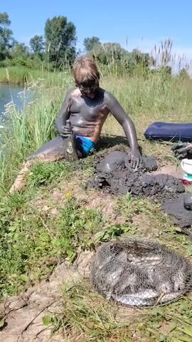 A young boy covered in mud sits on a grassy bank next to a body of water. He is holding a bottle and appears to be smearing mud on himself and playing in a pile of mud. A fishing net filled with fish is in the foreground. In the background, there are trees and a clear blue sky.