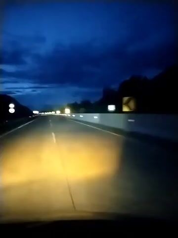 The video shows a first-person view from a vehicle traveling on a highway at night. The headlights illuminate the road ahead, casting a warm glow on the asphalt. The sky is a deep, dark blue, with clouds suggesting either twilight or a cloudy night. Lights are visible in the distance along the side of the highway, and silhouetted trees and possibly hills can be seen on the right side of the road. The highway appears to be empty, with no other vehicles clearly visible in the immediate vicinity. The white lines of the highway are prominent, guiding the path of travel. The overall impression is one of a solitary journey through the darkness.