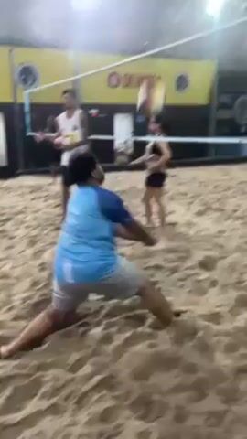 The video shows a beach volleyball game inside an indoor facility. Three people are visible: two men and one woman. The man in the foreground wears a light blue shirt and grey shorts. He dives to hit the volleyball. The woman in the background wears a red shirt, dark leggings, and is preparing to hit the volleyball. The other man wears a white tank top and black shorts. He is also preparing to hit the ball. A volleyball net separates the two sides. The ground is covered in sand. Bright lights illuminate the indoor space. Yellow advertising banners hang in the background.