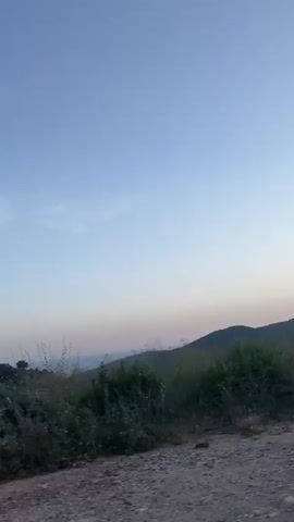 A video taken from a vehicle moving along a dirt road. The scene is rural, with rolling hills in the distance under a clear blue sky that is transitioning to a soft pink and orange hue near the horizon, indicating either sunrise or sunset. The immediate foreground consists of the dirt road and a dense growth of tall, green grass and weeds. A large, light-colored rock is visible on the left side of the road. Further back, more lush vegetation and a few scattered trees can be seen, with the landscape sloping upwards towards the hills. The overall mood is peaceful and serene, capturing the natural beauty of the landscape at a transitional time of day. There is no visible text in the video.