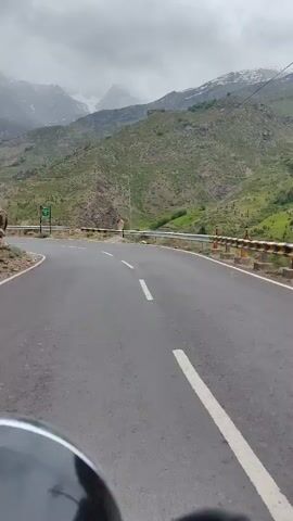A POV shot from a vehicle shows a winding mountain road under a cloudy sky. The road is asphalt with white lane markings. On the left side of the road, there are steep, green, and rocky hills with some snow-capped mountains visible in the distance. A green sign with white text and a railing are visible on the left. On the right, there is a guardrail. The lower part of the frame shows the dashboard and handlebars of the vehicle, suggesting it is a motorcycle. The camera pans slightly, following the curve of the road.
