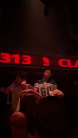 The video shows a dark interior with a neon red sign that reads "CLASS 313" in large block letters. Below the sign, three young men are visible. The man in the center wears a white and blue jersey with the number "86" prominently displayed. He appears to be talking or singing, as his mouth is open and he is gesturing with his hands. To his left, another man in a white t-shirt is also visible. The third man is on the right, mostly obscured by shadows and other people. The overall atmosphere suggests a nightclub or a similar entertainment venue.