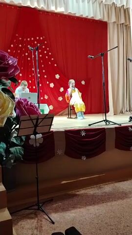 A stage is set up for a performance. A young girl in a colorful outfit sits on a chair in the center of the stage and sings into a microphone. Behind her is a red curtain decorated with white flowers. To her left, a young man sits at a table with a laptop and other equipment. In front of the stage are musical instrument stands and stage decoration. The stage is lit and the overall scene appears to be indoors in a public space.
