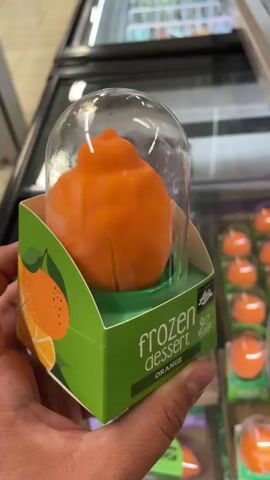 A person's hand holds a green cardboard box with a clear plastic dome containing an orange frozen dessert. The box reads "frozen dessert" and "ORANGE" in white text. A logo for "FRIENDS OF ASIA" and the word "VEGAN" are also visible on the box. The dessert itself is orange and appears to be shaped like a tangerine. The background shows a freezer aisle with more of these desserts and other frozen food items.