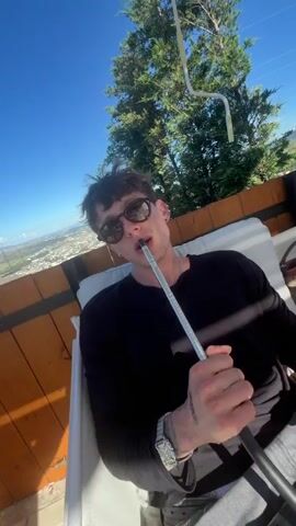 A man wearing sunglasses and a black long-sleeved shirt is sitting outdoors and smoking a hookah. The background shows a sunny day with a clear blue sky and trees. Another clip shows a group of people sitting at a table outdoors with drinks and a hookah. The scene appears to be a restaurant or lounge area. A third clip shows a sunny outdoor space with people relaxing on the grass and beanbag chairs. A building with "LOVE GARDENS" visible is in the background. The final clip shows a man in sunglasses taking a selfie with a scenic view of hills and a body of water in the background. He is sitting in an outdoor seating area with wooden accents. Several cars are parked in the background, including a black Tesla.