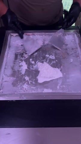 A person wearing black gloves and a light green t-shirt is preparing rolled ice cream on a metal cold plate. The person uses two metal spatulas to mix and spread a white creamy substance across the cold plate. The plate has a frosted appearance with remnants of previous mixing. The bottom of the plate is housed in a black unit.