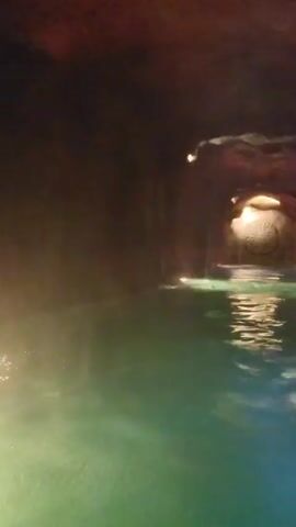 The video shows a narrow tunnel of artificial stone partially filled with water. The water is green. The walls of the tunnel are brown and uneven. The tunnel is dimly lit with small lights. There is steam rising from the water. At the far end of the tunnel there is a decorative stone carving in the wall. There is a hole in the ceiling of the tunnel that allows natural light to enter the space. There is talking in the background.