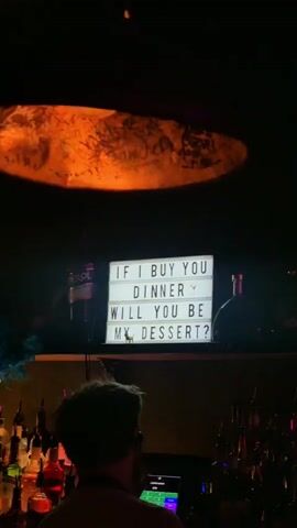 The video begins in a dimly lit bar. A lightbox sign displaying "If I buy you dinner, will you be my dessert?" is illuminated. A couple of people are silhouetted in front of the bar, with liquor bottles visible on the shelves. There's a textured light fixture hanging above. The camera pans to the right, revealing a wall decoration: an framed portrait of a leopard wearing a Renaissance-era collar and decorative clothing. The lighting shifts, highlighting the leopard's face in purple hues. The video ends with a close-up of the portrait.