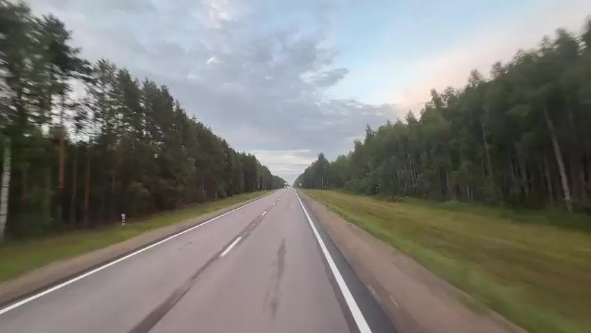 The video shows a point-of-view shot of a person driving on a two-lane asphalt road. The road is surrounded by a dense forest of pine trees on both sides. The sky is cloudy with some patches of blue. There is a white line marking the edge of the road and a double white line in the middle. Two trucks are visible in the distance, driving in the same direction as the camera. The road appears wet in places, possibly from recent rain. The camera is moving forward, creating a sense of motion blur. The time of day appears to be either early morning or late evening due to the soft lighting. There are no other people visible in the frame.