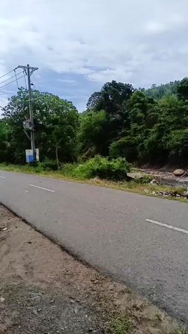 The camera pans to the left along a cracked, gray asphalt road next to a rocky riverbed. The river flows on the right side of the video, with green trees and hills in the background. A small structure is visible further down the river.