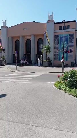 The video shows a street scene in front of a building with columns. The building has signs that read "THERMAL STRANDBAD" and a banner with "100 JAHRE". There are people entering and exiting the building. In the foreground, there is a crosswalk and a street. A green and yellow van drives by. Further into the video, a child is seen interacting with a bicycle that is parked next to a stone pillar. The child is touching the handlebars and the bell of the bicycle. The environment appears to be outdoors on a sunny day.