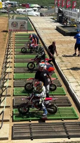 The video shows a motocross starting line with several young riders on their bikes, waiting for the signal to begin the race. The riders are lined up on individual metal grids, each wearing helmets and protective gear. Several adults are around, appearing to be coaches or parents. The background shows a racetrack with tents, flags, and spectators. The day appears sunny, with clear visibility.
