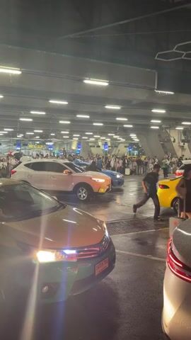 The video shows a crowded outdoor area, likely a taxi stand or pick-up zone at an airport or large venue. Several cars are visible, including a green taxi in the foreground, a white SUV, a pink SUV, a blue sedan, and a yellow taxi. A large number of people are standing and walking in the background, many with luggage, suggesting they are travelers. The lighting appears to be artificial, from overhead fixtures, and the ground is wet, indicating recent rain. The overall atmosphere seems busy and somewhat chaotic.