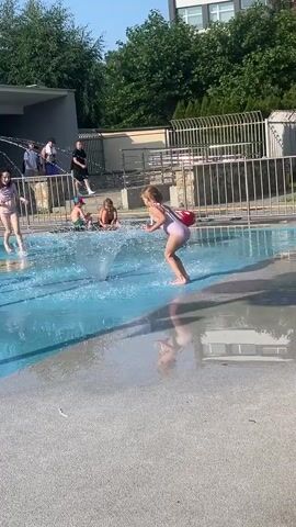 A girl in a swimsuit jumps through a fountain in a shallow pool. Other children play in the water behind her. People stand near the edge of the pool. Trees and a building are visible in the background.