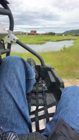 The video opens from the perspective of someone riding in a side-by-side vehicle. The camera is pointed forward, showing the legs and feet of the rider in blue jeans and patterned sneakers. The vehicle's interior, including a black net seatbelt and a metal floor, is visible. Beyond the vehicle, a rural landscape unfolds under a cloudy sky. A body of water, possibly a lake or river, is bordered by lush green grass and scattered buildings. In the distance, houses and other structures are visible. The vehicle appears to be moving along a dirt or gravel path. The camera then pans to show the wider environment, revealing more of the rural settlement, with several small buildings lined up along a dirt road. A body of water is visible to the left, with reeds and tall grass growing near its edge. The video continues to show the vehicle traveling through the grassy terrain, with the landscape of scattered houses, water, and fields remaining in view. The sky is overcast with thick clouds throughout the duration of the video.