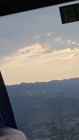 The video captures a view from a moving vehicle, likely a bus, looking out a window. The foreground shows a portion of the vehicle's interior, including dark fabric and a sticker with a running figure symbol and Chinese characters. The middle ground reveals a vast mountainous landscape with a prominent electricity pylon and power lines stretching across the scene. In the distance, a city with high-rise buildings is visible nestled among the hills, and a bus is seen driving on a road in the lower left corner. The sky is partly cloudy with a gradient of blue and soft yellow hues, suggesting it might be dawn or dusk. The overall impression is one of a scenic journey through a developed mountainous region.