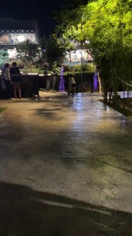 The media file depicts an outdoor scene at night. In the foreground, a wet, textured concrete pathway reflects the ambient light, suggesting it might have rained or been recently cleaned. On the left, three individuals are partially visible, standing near a dark railing. One person appears to be wearing a dark t-shirt and shorts, another a light-colored top, and the third is obscured.

Further into the scene, on the pathway, a lone figure, possibly a child or a young person wearing a light-colored hoodie, is interacting with something on the ground. Behind this figure, a black metal railing runs horizontally, separating the pathway from a landscaped area. Beyond the railing, a dark water feature with purple uplighting adds a splash of color to the night.

In the background, several modern buildings are visible, illuminated by interior lights. The architecture suggests a commercial or entertainment complex. Lush green trees, some appearing to be bamboo, frame the right side of the image, their leaves catching the light.

The overall impression is of a lively urban setting at night, possibly a park, a promenade, or an outdoor dining area in Davao City, Philippines. The time of day is clearly night due to the artificial lighting and darkness. The wet pathway implies recent rain or cleaning. No text is visible in the media file. The mood seems relaxed and casual, with people enjoying the evening atmosphere.
