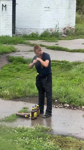 A person in a dark blue shirt and black pants bends over and picks up a brown bottle with a yellow cap. They bring the bottle to their face and appear to smell it. The person is standing on a wet, paved surface surrounded by grass. In front of them, there are five similar bottles standing upright in a line, and two brown cylindrical objects are lying on the ground. Behind the person is a white brick building with graffiti on it.