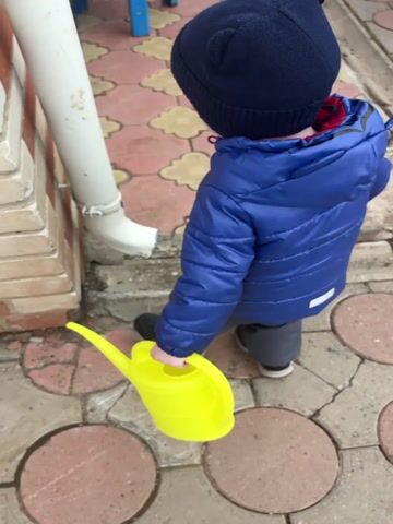 A young child, viewed from behind and slightly above, is standing outdoors. The child is dressed in a dark blue knit beanie with small ear-like protrusions, a bright blue quilted winter jacket, dark grey pants, and dark shoes with white soles. They are holding a bright yellow plastic watering can in their right hand.

The child is positioned next to a brick wall, which has a white downspout running vertically down its side. The watering can is held with its spout pointed towards the downspout, suggesting the child is attempting to collect water from it. The ground beneath the child is paved with a pattern of reddish-brown and light grey pavers, some of which are circular and some resemble cloud shapes.

In the background, more of the paved surface is visible, along with parts of the brick wall. The lighting suggests it is daytime, possibly overcast.

A child's voice is heard saying "Водичка иди, иди, иди" (Vodichka idi, idi, idi), which translates to "Water come, come, come," and then "иди" (idi - "come"). An adult's voice is also heard saying "Ой" (Oy), which means "Oh" or "Oops."