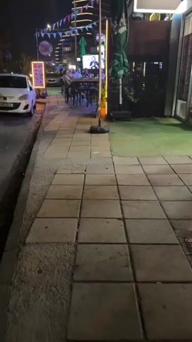 The video shows a nighttime street scene with a sidewalk in the foreground. To the left, a white car is parked on the street. The sidewalk is paved with large square tiles, some of which appear cracked or uneven. A small patch of gravel is visible next to the sidewalk. In the background, there is an outdoor seating area of a restaurant or cafe. Several people are seated at tables, some of whom are visible on a large screen in the background. The area is illuminated by various lights, including string lights, neon signs, and streetlights. A green umbrella is visible behind the seating area. The overall environment suggests an urban setting at night.