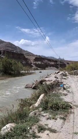 The video shows a scenic landscape with a winding road. On the left side of the road, there is a river with flowing water and rocky banks. Mountains form the backdrop, with some sparse vegetation along the river and the mountainsides. Power lines are visible crossing the sky. The road itself is paved with white lines marking the lanes. The sky is blue with scattered white clouds. The scene appears to be in a mountainous or desert region, with clear weather.